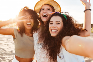 Three girls taking a beachside selfie, laughing and smiling in the sun